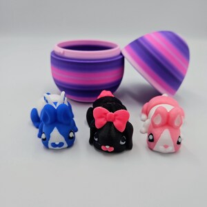 K&ouml;nnte beinhalten: Drei kleine Pl&uuml;sch-Hasenspielzeuge in einem lila und pink gestreiften eif&ouml;rmigen Beh&auml;lter. Die Hasen sind blau, schwarz mit einer rosa Schleife und pink.