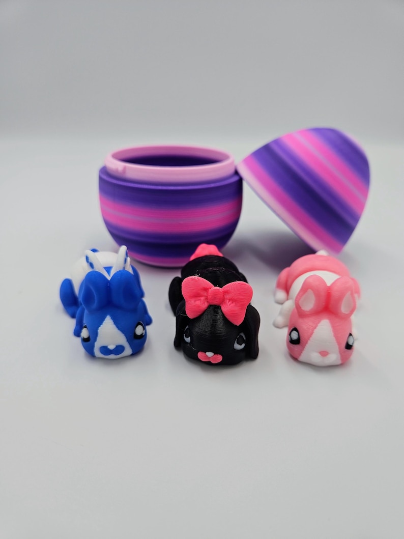 K&ouml;nnte beinhalten: Drei kleine Pl&uuml;sch-Hasenspielzeuge in einem lila und pink gestreiften eif&ouml;rmigen Beh&auml;lter. Die Hasen sind blau, schwarz mit einer rosa Schleife und pink.