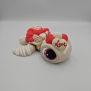 Pode incluir: Uma figura de tartaruga bege e caprichosa com um olho grande e uma concha decorada em vermelho e branco. A concha &eacute; adornada com cora&ccedil;&otilde;es vermelhos, doces brancos e a palavra "Love" em escrita branca. As barbatanas da tartaruga s&atilde;o bege com listras brancas.