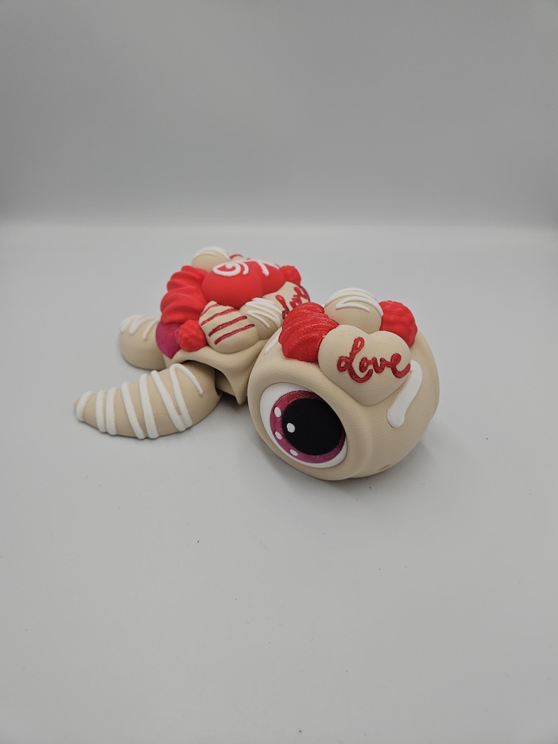 Pode incluir: Uma figura de tartaruga bege e caprichosa com um olho grande e uma concha decorada em vermelho e branco. A concha &eacute; adornada com cora&ccedil;&otilde;es vermelhos, doces brancos e a palavra "Love" em escrita branca. As barbatanas da tartaruga s&atilde;o bege com listras brancas.
