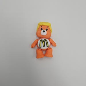 May include: An orange bear-like figurine with a yellow hairstyle and a white belly featuring a yellow and black "M" logo. The figurine has black eyes, orange arms, and orange feet. The overall design is a playful, cartoonish style.