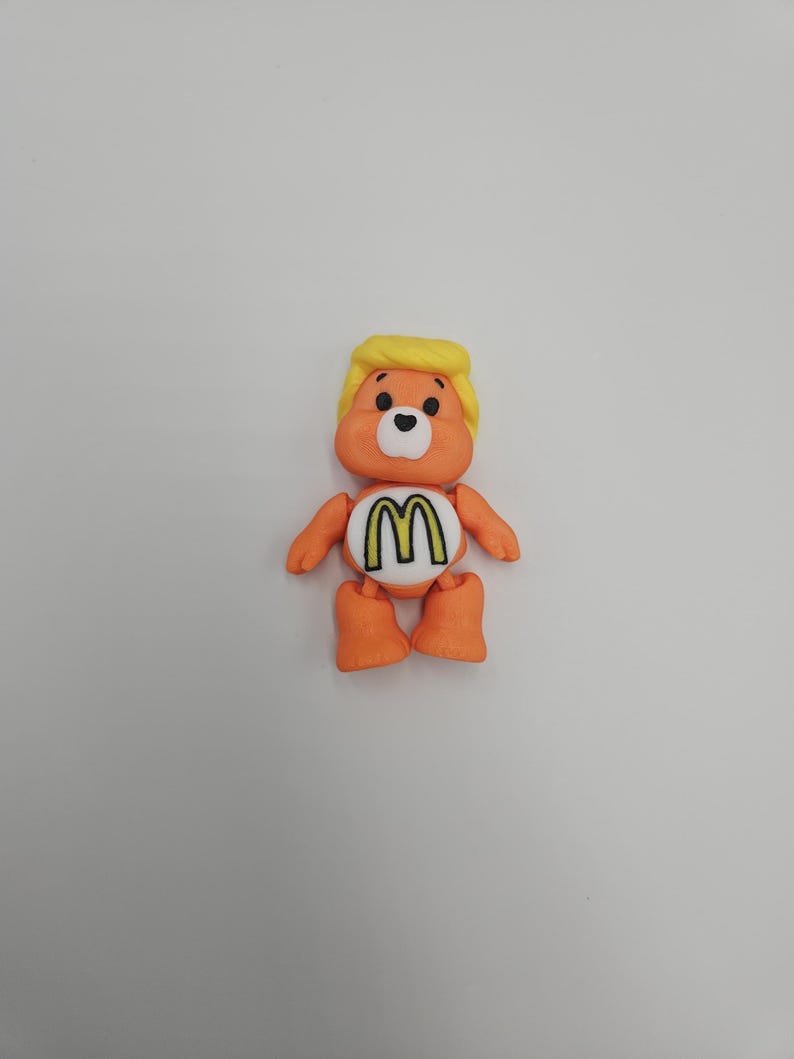May include: An orange bear-like figurine with a yellow hairstyle and a white belly featuring a yellow and black "M" logo. The figurine has black eyes, orange arms, and orange feet. The overall design is a playful, cartoonish style.
