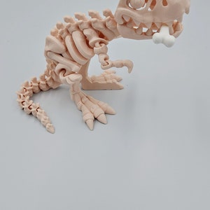 May include: A light pink plastic dinosaur skeleton toy with a bone in its mouth. The dinosaur is posed in a standing position with its tail curled behind it.