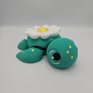 Pode incluir: Uma figura de tartaruga turquesa caprichosa com uma flor de margarida branca e amarela nas costas. A tartaruga tem manchas amarelas e olhos grandes e expressivos. Este item decorativo &eacute; provavelmente uma pe&ccedil;a artesanal.