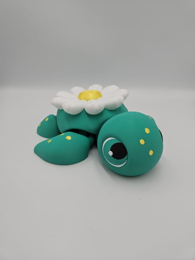 Pode incluir: Uma figura de tartaruga turquesa caprichosa com uma flor de margarida branca e amarela nas costas. A tartaruga tem manchas amarelas e olhos grandes e expressivos. Este item decorativo &eacute; provavelmente uma pe&ccedil;a artesanal.