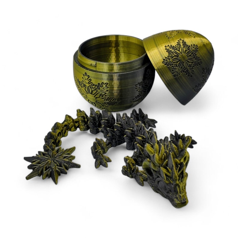 K&ouml;nnte beinhalten: Ein schwarzes und goldenes 3D-gedrucktes Drachenpuzzle. Der Drache besteht aus vielen kleinen Teilen, die ineinanderpassen. Der Drache sitzt auf einem wei&szlig;en Hintergrund. Der Drache befindet sich in einem geschlossenen, goldenen, eif&ouml;rmigen Beh&auml;lter mit einem Schneeflockenmuster.