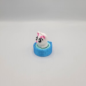 May include: A white cat figurine with pink accents, including ears and spots, sits atop a light blue well-shaped base. The cat has large black eyes. The base is a darker blue with a brick pattern.