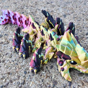 May include: A multi-colored, iridescent, 3D printed dragon figurine with a scaly texture. The dragon is curled up with its head resting on the ground. The dragon is painted in shades of pink, yellow, green, and black.