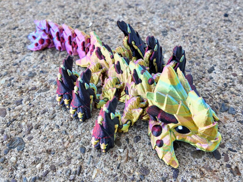 May include: A multi-colored, iridescent, 3D printed dragon figurine with a scaly texture. The dragon is curled up with its head resting on the ground. The dragon is painted in shades of pink, yellow, green, and black.