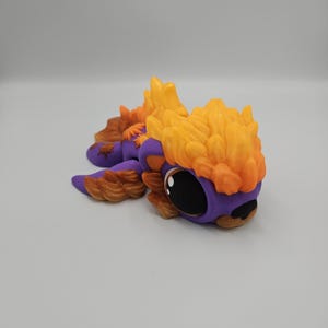Pode incluir: Figurinha artesanal e caprichosa de um pequeno drag&atilde;o. O drag&atilde;o apresenta um corpo roxo, olhos pretos grandes e detalhes em laranja e castanho. A cabe&ccedil;a e as costas do drag&atilde;o s&atilde;o adornadas com detalhes texturizados em forma de chama.
