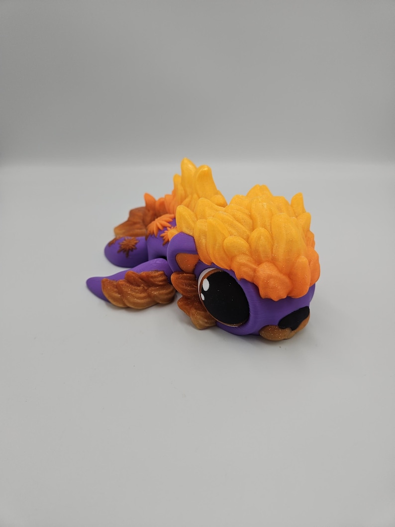 Pode incluir: Figurinha artesanal e caprichosa de um pequeno drag&atilde;o. O drag&atilde;o apresenta um corpo roxo, olhos pretos grandes e detalhes em laranja e castanho. A cabe&ccedil;a e as costas do drag&atilde;o s&atilde;o adornadas com detalhes texturizados em forma de chama.