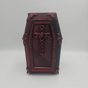 May include: A burgundy-colored coffin-shaped box with ornate detailing. The box features a cross design on the front and decorative trim around the edges. The coffin has a textured surface and is set against a plain background.