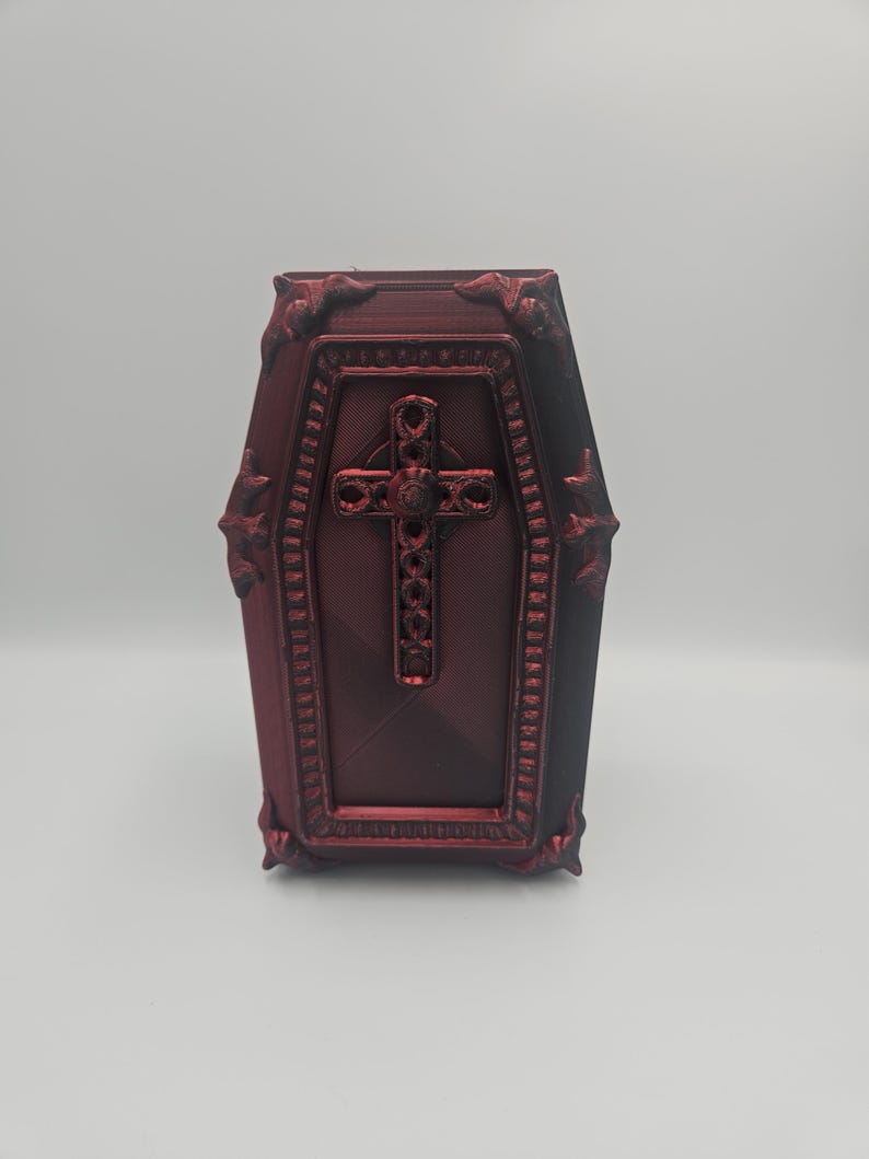 May include: A burgundy-colored coffin-shaped box with ornate detailing. The box features a cross design on the front and decorative trim around the edges. The coffin has a textured surface and is set against a plain background.