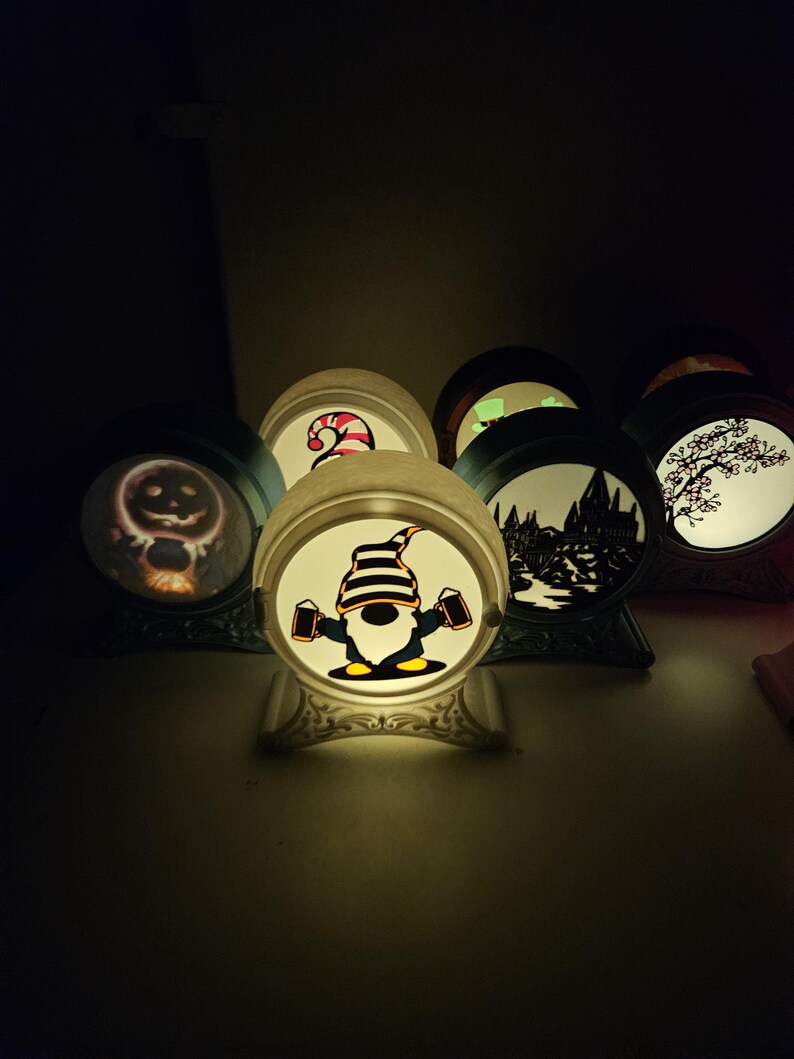 May include: A collection of illuminated decorative globes with various designs. One globe features a gnome holding beer steins, while others display a jack-o'-lantern, a candy cane, a silhouette of a castle, and a tree. The globes have ornate bases.