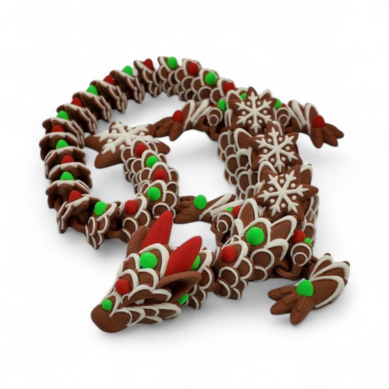 Puede incluir: Un caprichoso drag&oacute;n de pan de jengibre, decorado con glaseado blanco, caramelos rojos y verdes y detalles de copos de nieve. El cuerpo del drag&oacute;n est&aacute; hecho de piezas de pan de jengibre interconectadas, una decoraci&oacute;n navide&ntilde;a &uacute;nica.