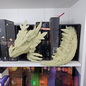 Op de afbeelding: Een paar lichtgroene boekensteunen in de vorm van een draak. De boekensteunen zijn gemaakt van een textuurrijk materiaal en zijn ontworpen om boeken op een plank te houden. De drakenkop en -staart zijn gedetailleerd.