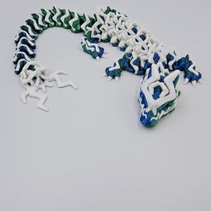 May include: A 3D printed lizard figurine with a white and green segmented body and a blue and green head. The lizard is posed in a curled position with its tail curled around its body.