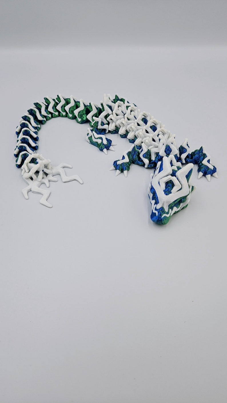 May include: A 3D printed lizard figurine with a white and green segmented body and a blue and green head. The lizard is posed in a curled position with its tail curled around its body.
