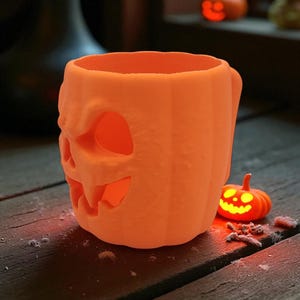 Pode incluir: Caneca laranja com tema de Halloween em forma de ab&oacute;bora esculpida com uma al&ccedil;a. A ab&oacute;bora tem um rosto esculpido com olhos e boca brilhantes. Uma pequena ab&oacute;bora brilhante est&aacute; ao lado da caneca.