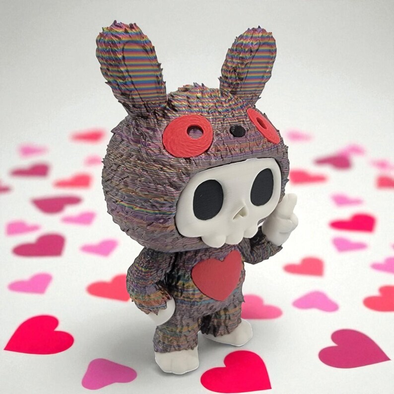 K&ouml;nnte beinhalten: Eine einzigartige Figur eines Hasen mit Totenkopfmaske und Herz auf der Brust. Der Hase ist &uuml;berwiegend dunkel mit bunten Streifen, roten Akzenten und wei&szlig;en Details. Er steht auf einer Oberfl&auml;che, die mit rosa und roten Herzformen &uuml;bers&auml;t ist.