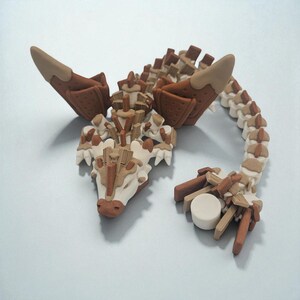 以下が含まれることがあります： 茶色、ベージュ、クリーム色の手作りのドラゴン彫刻。ドラゴンは、詳細な翼、セグメント化された体、様式化された頭部が特徴です。彫刻は、さまざまな幾何学的な形とテクスチャで作られています。
