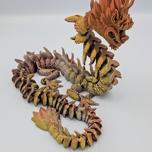 May include: A detailed, articulated dragon figurine in shades of gold, brown, and orange. The dragon has a detailed head, spiky mane, and segmented body. The figurine is posed on a white surface.