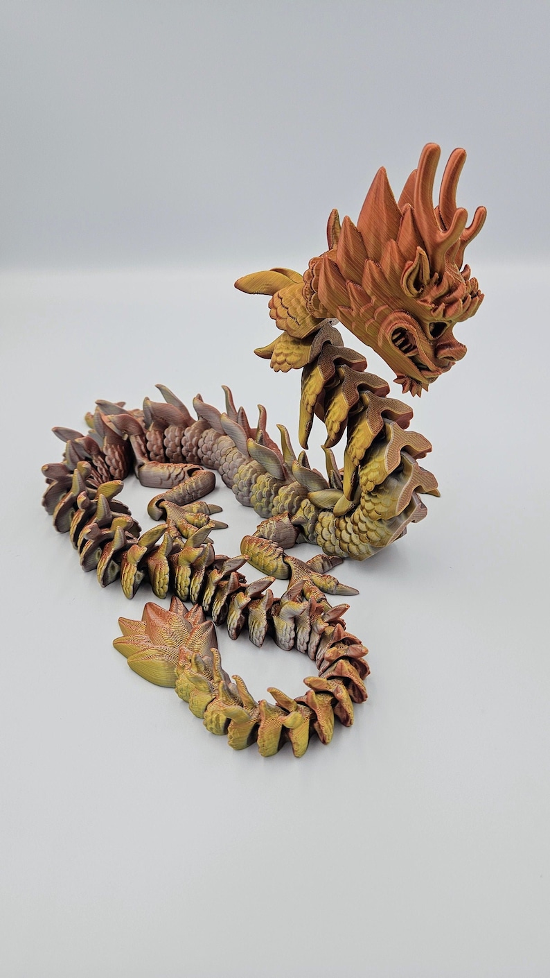 May include: A detailed, articulated dragon figurine in shades of gold, brown, and orange. The dragon has a detailed head, spiky mane, and segmented body. The figurine is posed on a white surface.