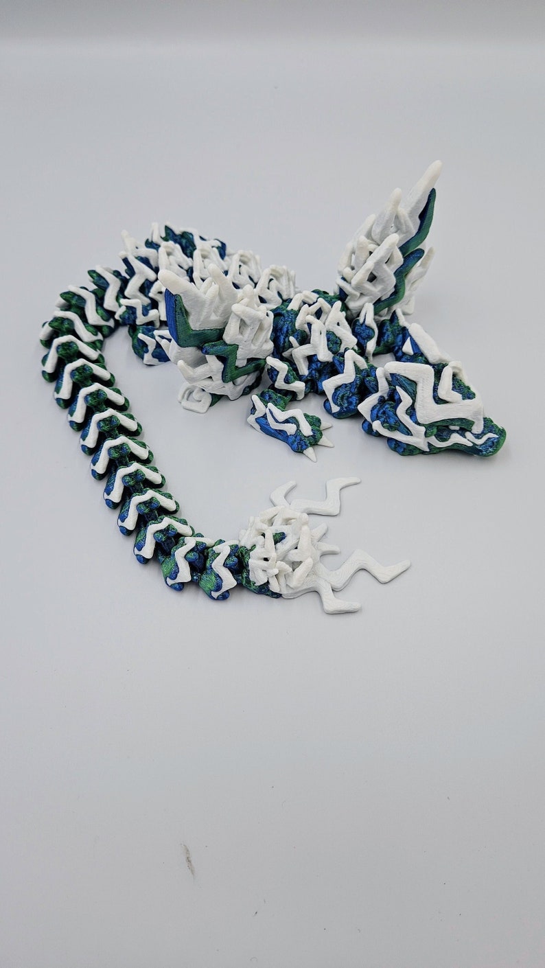 May include: A 3D printed dragon figurine with a white and green colour scheme. The dragon has a long, segmented tail and a detailed head with sharp teeth.