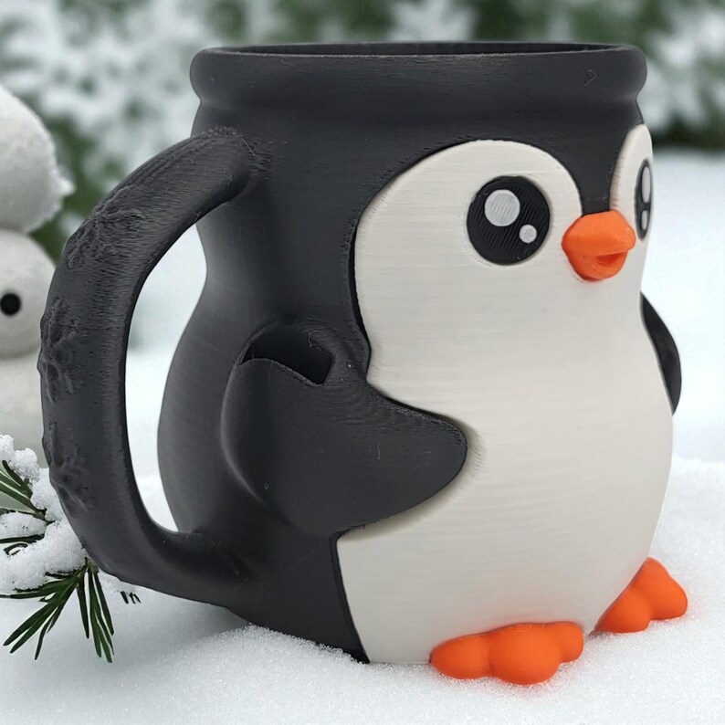 May include: A black, white, and orange penguin-shaped mug. The mug features a detailed handle with a snowflake design. The penguin has large eyes and orange feet and beak. The mug is sitting in the snow.