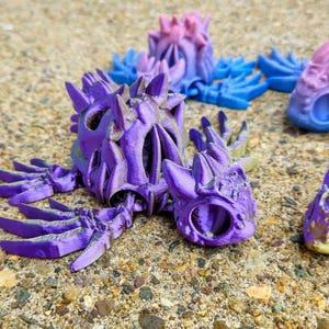 Peut inclure: Figurines de dragon imprim&eacute;es en 3D de couleur violette, bleue et rose avec des d&eacute;tails complexes et un design squelettique. Les dragons sont pos&eacute;s dans diff&eacute;rentes positions, les ailes d&eacute;ploy&eacute;es et les queues enroul&eacute;es.