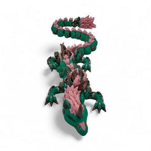 May include: A 3D-printed dragon figurine in a vibrant green and pink color scheme. The dragon has a detailed, textured body with a serpentine form, featuring spiky protrusions along its back and head. The dragon's claws and horns are a darker shade, adding contrast.