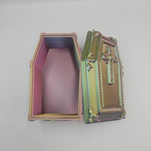 May include: An open, iridescent coffin-shaped box with a hinged lid. The box is painted with a gradient of colors, including pink, yellow, and green. The lid features a cross and decorative embellishments. The interior of the box is a solid pink color.