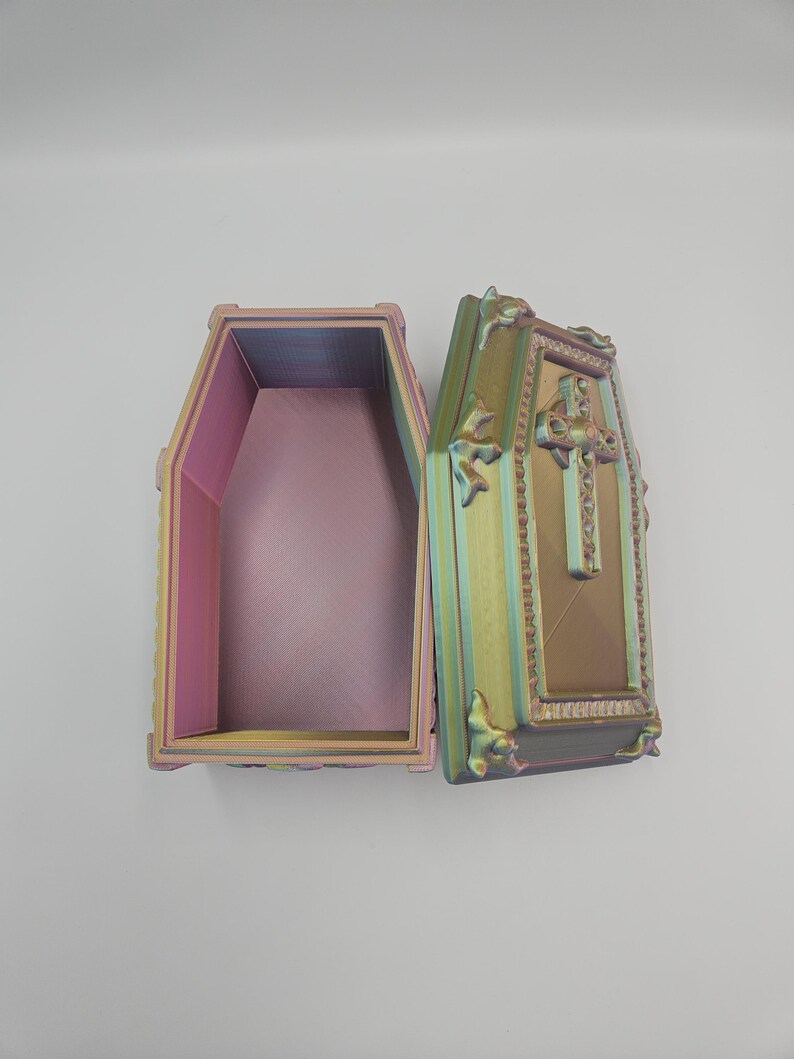 May include: An open, iridescent coffin-shaped box with a hinged lid. The box is painted with a gradient of colors, including pink, yellow, and green. The lid features a cross and decorative embellishments. The interior of the box is a solid pink color.