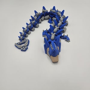 Pode incluir: Um brinquedo de drag&atilde;o articulado azul e cinza com o texto "USAF" e "AIR FORCE". O drag&atilde;o &eacute; feito de pl&aacute;stico e tem um design detalhado, com espinhos e um corpo segmentado. O brinquedo est&aacute; posicionado em uma superf&iacute;cie branca.