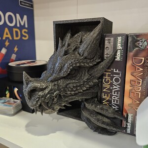 May include: A black dragon head bookend with a textured, glittery finish. The bookend is holding up two board game boxes, one titled "One Night Werewolf" and the other titled "Daybreak".