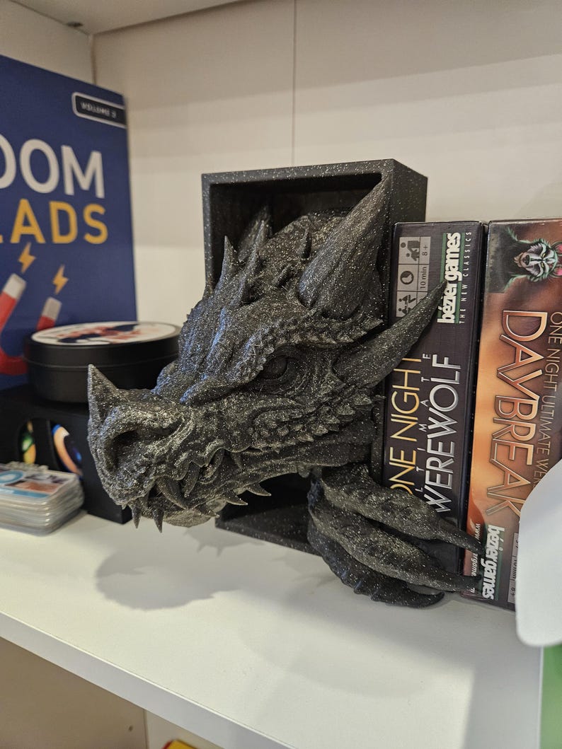 May include: A black dragon head bookend with a textured, glittery finish. The bookend is holding up two board game boxes, one titled "One Night Werewolf" and the other titled "Daybreak".