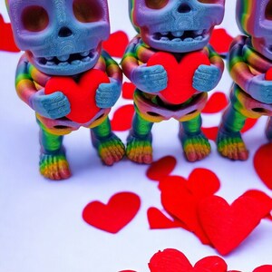May include: Three rainbow-coloured skeleton figurines holding red hearts. The skeletons are standing on a white surface with red paper hearts scattered around them.