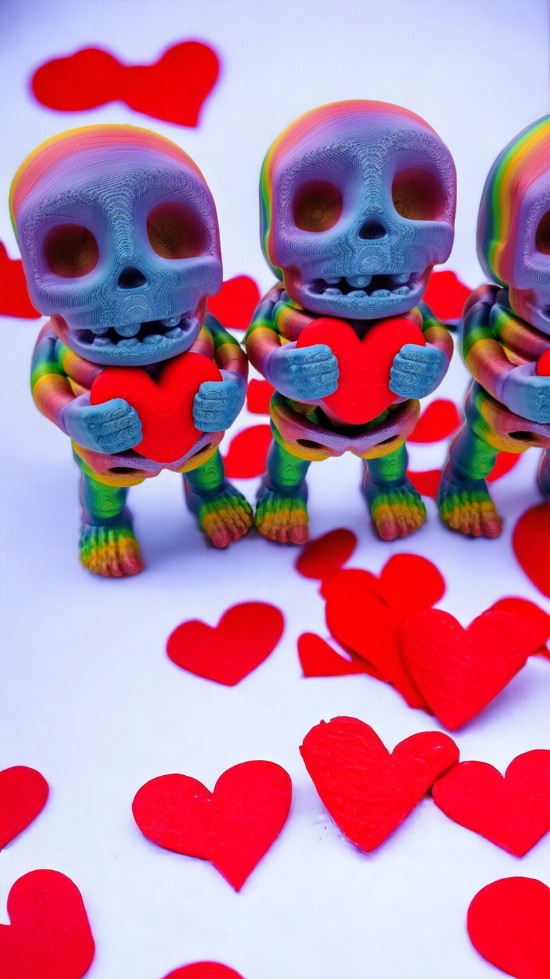 May include: Three rainbow-coloured skeleton figurines holding red hearts. The skeletons are standing on a white surface with red paper hearts scattered around them.