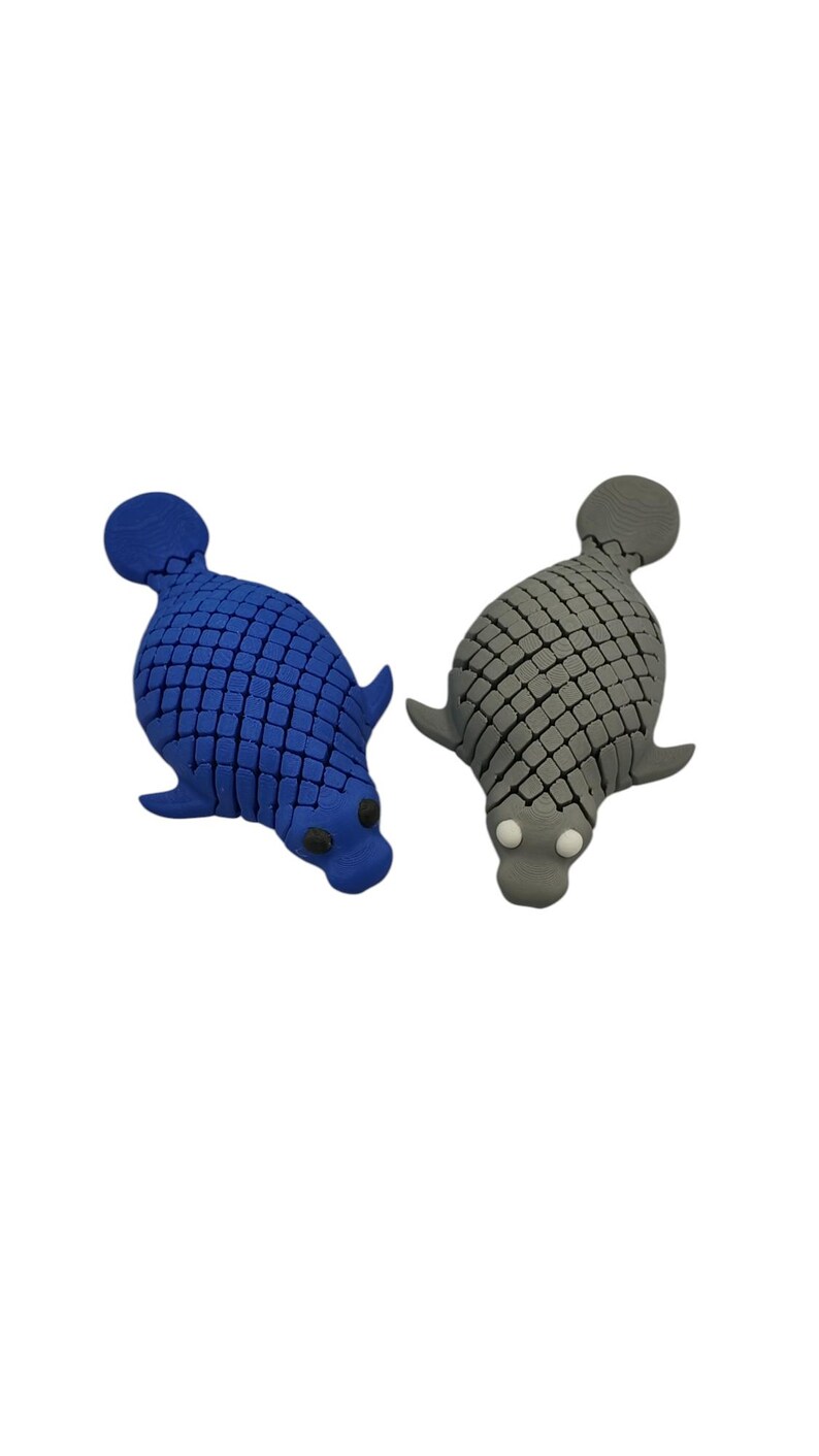 Puede incluir: Dos juguetes con forma de foca. Uno es azul con un patr&oacute;n de cuadr&iacute;cula, el otro es gris con un patr&oacute;n similar. Ambos tienen aletas, una cola y una cabeza con ojos peque&ntilde;os y oscuros. Los juguetes est&aacute;n sobre un fondo blanco.
