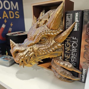 May include: A gold dragon head bookend with a detailed design. The bookend is holding up a book titled "One Night Werewolf" by Bezier Games.