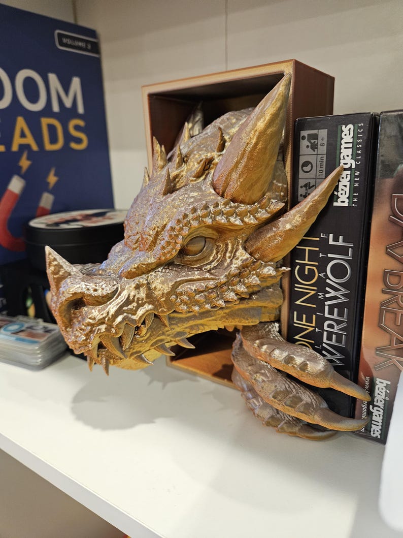 May include: A gold dragon head bookend with a detailed design. The bookend is holding up a book titled "One Night Werewolf" by Bezier Games.