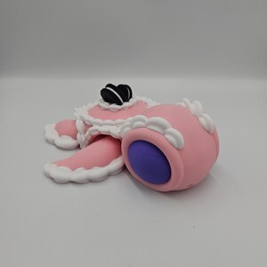 Pode incluir: Uma escultura rosa caprichosa que se assemelha a uma sobremesa, com um corpo redondo e um interior roxo. Apresenta um topo decorativo com detalhes em preto e branco e acabamento branco. O design &eacute; caricatural e l&uacute;dico.