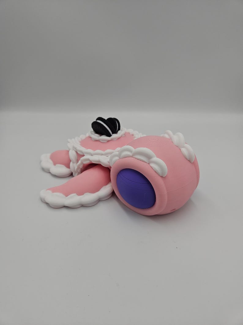 Pode incluir: Uma escultura rosa caprichosa que se assemelha a uma sobremesa, com um corpo redondo e um interior roxo. Apresenta um topo decorativo com detalhes em preto e branco e acabamento branco. O design &eacute; caricatural e l&uacute;dico.