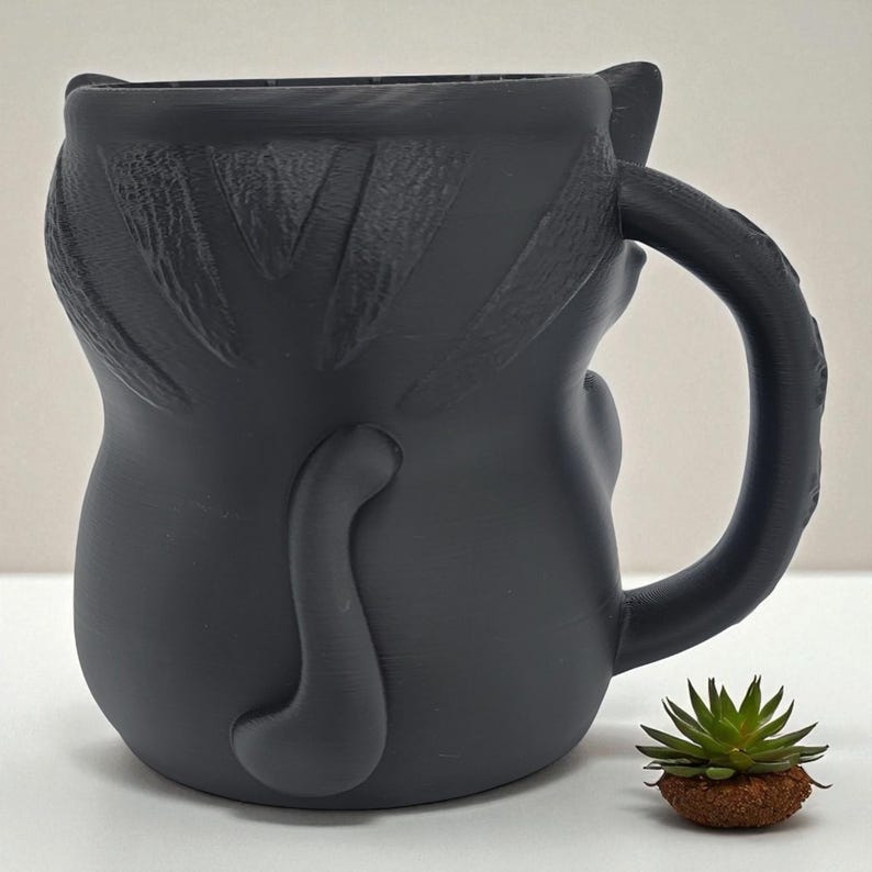 May include: A matte black ceramic mug shaped like a cat, featuring a handle, ears, and a tail. The mug has textured details and a smooth finish. A small succulent plant sits next to the mug.