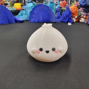Peut inclure: Figurine blanche imprim&eacute;e en 3D en forme de ravioli, avec un visage souriant. La figurine a des yeux noirs, une bouche noire et des joues roses. D'autres objets imprim&eacute;s en 3D color&eacute;s sont visibles en arri&egrave;re-plan.