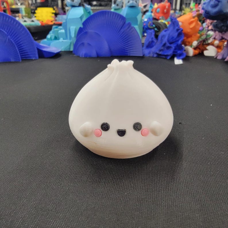 Peut inclure: Figurine blanche imprim&eacute;e en 3D en forme de ravioli, avec un visage souriant. La figurine a des yeux noirs, une bouche noire et des joues roses. D'autres objets imprim&eacute;s en 3D color&eacute;s sont visibles en arri&egrave;re-plan.