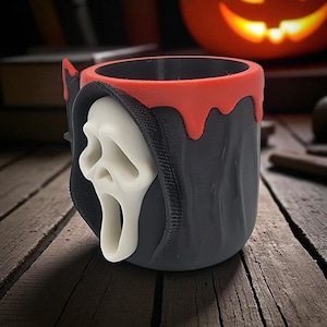 Pode incluir: Uma caneca preta e vermelha com tema de Halloween, com uma m&aacute;scara Ghostface branca. A caneca tem uma borda vermelha e um design de capa preta. O design &eacute; impresso em 3D e fica em uma superf&iacute;cie de madeira.