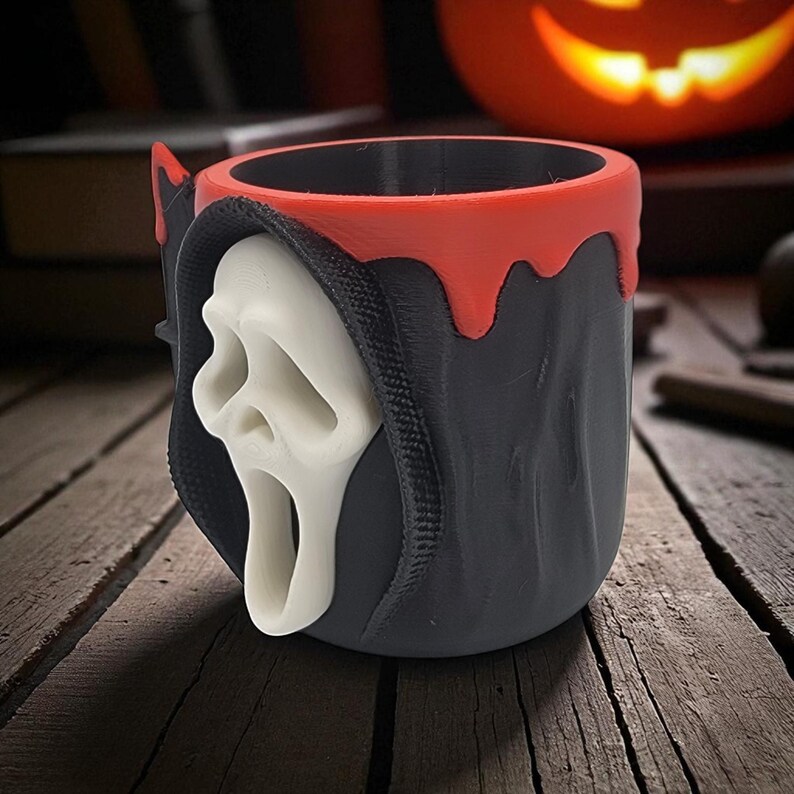 Pode incluir: Uma caneca preta e vermelha com tema de Halloween, com uma m&aacute;scara Ghostface branca. A caneca tem uma borda vermelha e um design de capa preta. O design &eacute; impresso em 3D e fica em uma superf&iacute;cie de madeira.
