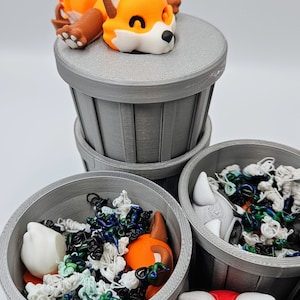 May include: A collection of gray trash cans, each containing colorful, abstract objects and animal figurines. A sleeping orange fox figurine rests atop one can. Other figurines include a raccoon and a red fox.