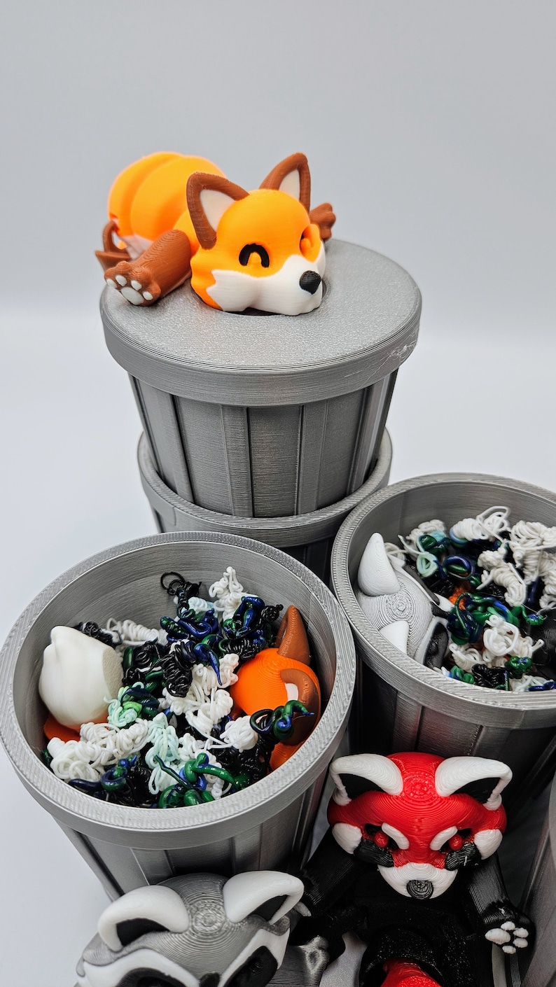 May include: A collection of gray trash cans, each containing colorful, abstract objects and animal figurines. A sleeping orange fox figurine rests atop one can. Other figurines include a raccoon and a red fox.